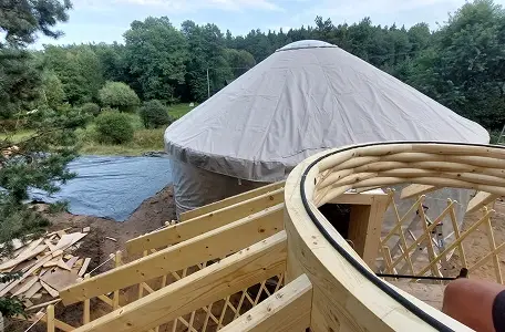 Budowa jurty, etap 4: Widok na drewnianą kratownicę dachową w trakcie montażu, w tle widoczny zielony las.